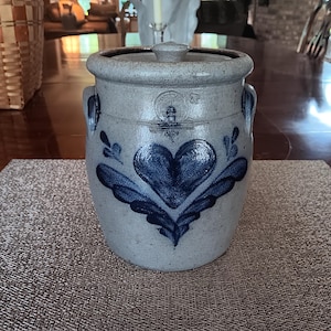May include: A stoneware crock with a lid, featuring a blue heart design and floral accents. The gray-toned jar has two handles and is likely used for food storage. The crock is sitting on a textured placemat.