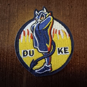 May include: Embroidered patch featuring a cartoon devil character in blue and red, with the word "DUKE" in yellow letters. The devil is surrounded by a yellow circle with flames.