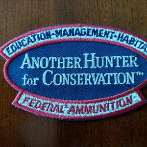 May include: A blue and red embroidered patch with the text "Another Hunter for Conservation" and "Federal Ammunition" in white. The patch also includes the text "Education-Management-Habitat" in white.