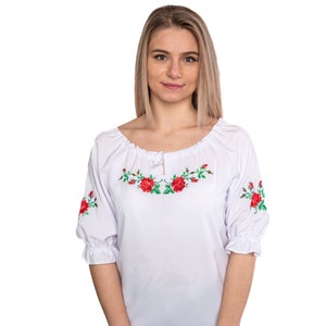 Puede incluir: Blusa blanca bordada con rosas rojas y hojas verdes. La blusa tiene un escote fruncido y mangas 3/4.