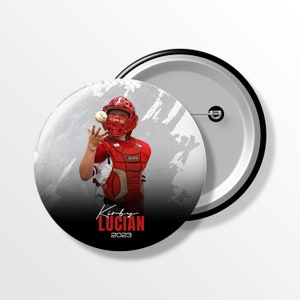 May include: A circular pinback button featuring a baseball catcher in red gear catching a ball. The design includes a white and gray textured background with the text Kirby LUCIAN 2023 printed in white and red at the bottom of the button face.