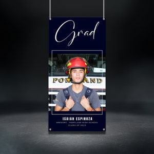 May include: A graduation banner with a navy blue background and white text that says "Grad". The banner features a photo of a young man wearing a fire helmet and a grey shirt. The text below the photo reads "ISAIAH ESPINOZA GREGORY - PORTLAND HIGH SCHOOL CLASS OF 2023".