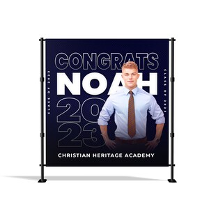 May include: A blue and white graduation banner with the text "CONGRATS NOAH 2023" and "CLASS OF 2023". A young man in a light blue shirt and a striped tie is standing in front of the banner.
