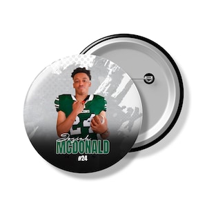 May include: A white and black button with a football player in a green jersey. The player is holding a football and has a black background with the text "Josiah MCDONALD #24".