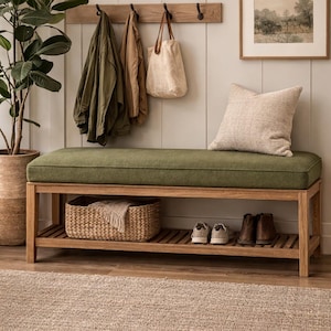 May include: A wooden bench with a green cushion, a lower shelf, and a woven basket. Above, a wooden rack holds coats and a tote bag. A framed picture and a decorative pillow complete the entryway scene.
