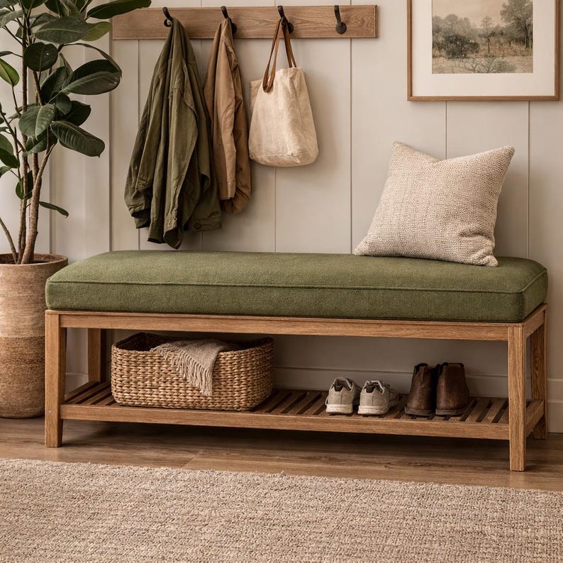 May include: A wooden bench with a green cushion, a lower shelf, and a woven basket. Above, a wooden rack holds coats and a tote bag. A framed picture and a decorative pillow complete the entryway scene.