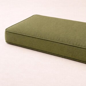 May include: Olive green rectangular cushion with a piped edge. The cushion has a textured fabric and a simple, modern design. The cushion is likely for a bench or outdoor seating.