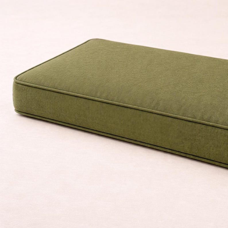 May include: Olive green rectangular cushion with a piped edge. The cushion has a textured fabric and a simple, modern design. The cushion is likely for a bench or outdoor seating.