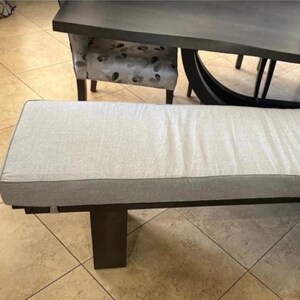 May include: A rectangular dining bench featuring a light gray upholstered seat cushion with a smooth, flat surface. The bench is supported by four sturdy, dark brown wooden legs that provide a stable base for the long, horizontal seating area.