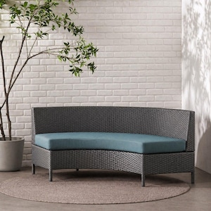 Könnte beinhalten: Ein graues Outdoor-Sofa aus Korbgeflecht mit geschwungener Rückenlehne und tealfarbenem Kissen. Das Sofa steht auf einem runden, neutralfarbenen Teppich. Ein Topfpflanze und eine weiße Backsteinmauer sind im Hintergrund.