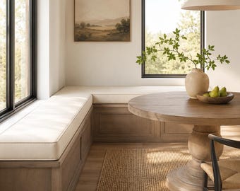 May include: A dining nook featuring a light wood built-in bench with a cream cushion, a round wooden pedestal table, and a black chair. A ceramic vase with green branches and a bowl of pears sit on the table, with a landscape painting on the wall.