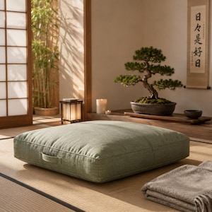 May include: A square, sage green floor cushion with a textured fabric finish and a small side handle. The cushion is low-profile and sits flat on a woven surface, featuring clean, straight edges and a simple, uniform construction throughout.