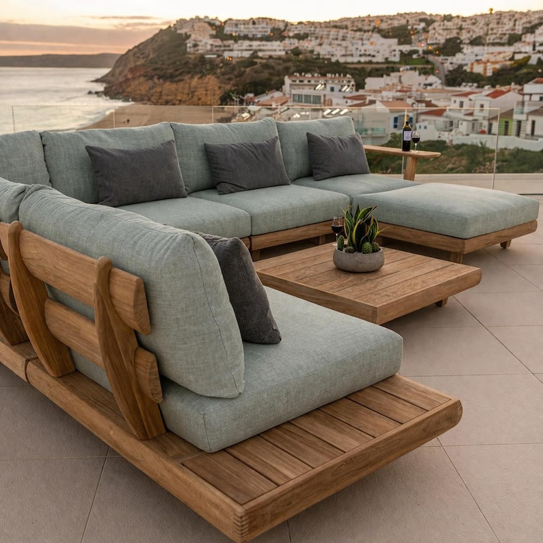 May include: A modular outdoor sectional sofa set with a natural teak wood frame and light sage green fabric cushions. The set includes a low-profile square coffee table and several dark gray rectangular throw pillows arranged on the seating.