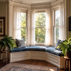 May include: A white built-in bay window seat with a dark blue cushion and two gray throw pillows. The seat is framed by white wooden trim and sheer white curtains. Two potted green ferns sit on small wooden stands on either side of the bench.