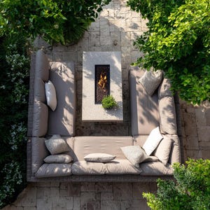 May include: An outdoor sectional sofa with taupe cushions and decorative pillows surrounds a rectangular coffee table with a built-in fire pit. The setting is surrounded by lush greenery and stone pavers.