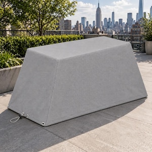 May include: A light gray rectangular protective cover for outdoor furniture, featuring a tapered shape with reinforced metal grommets at the bottom corners and an adjustable drawstring cord for a secure fit on a flat patio surface.