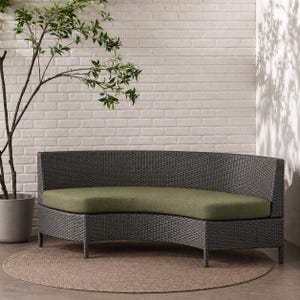 May include: A curved sofa featuring a dark gray woven wicker frame and a solid olive green seat cushion. The piece is supported by four thin, straight legs and has a continuous backrest that follows the arc of the seat frame design.