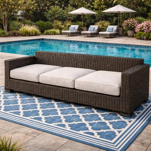 Op de afbeelding: Een donkerbruine wicker loungesofa met drie lichtgekleurde kussens. De bank staat op een blauw-wit patroonkleed. De achtergrond omvat een zwembad, ligstoelen en parasols.