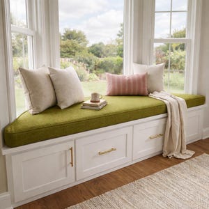 May include: A white built-in window seat with three storage drawers featuring gold handles, topped with a textured olive green cushion. Two beige square pillows, one pink striped rectangular pillow, a ceramic mug, and a throw blanket are included.