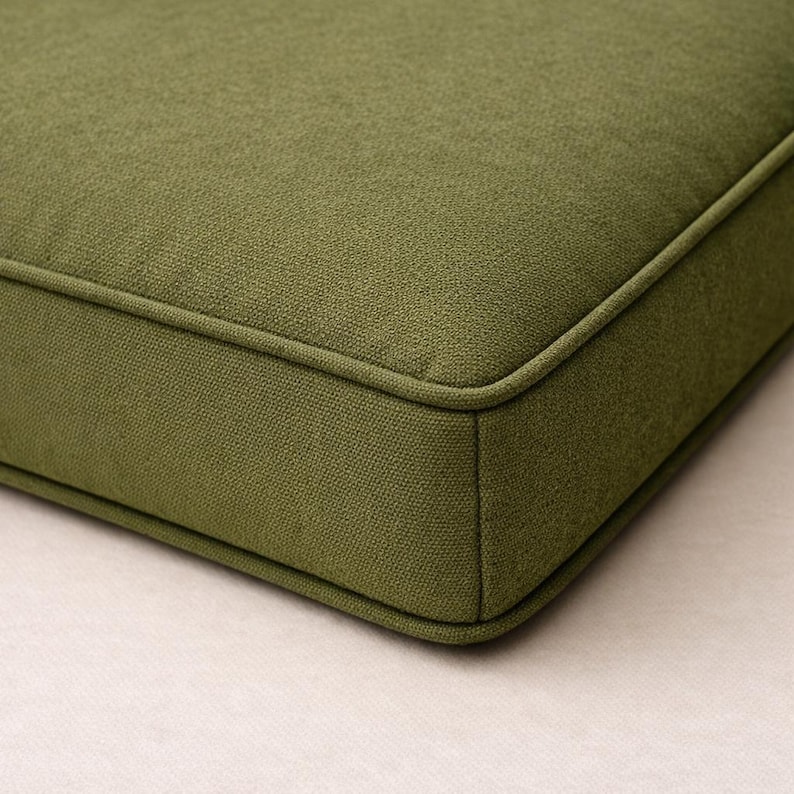 May include: Close-up of a square, olive-green cushion with a textured fabric. The cushion has a piped edge in a matching green color. The corner of the cushion is visible, showing the construction and the fabric's weave.