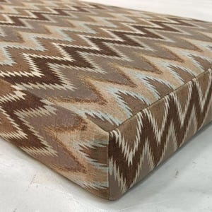 May include: Close-up of a rectangular cushion with a brown, tan, and light blue zigzag pattern. The cushion has a soft, textured appearance and a neutral color palette, suitable for various interior design styles.
