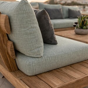 May include: A modular outdoor sofa section constructed from natural teak wood with a slatted base and frame. It features a thick, light sage green fabric seat cushion and a matching back cushion, complemented by a dark charcoal gray accent pillow.