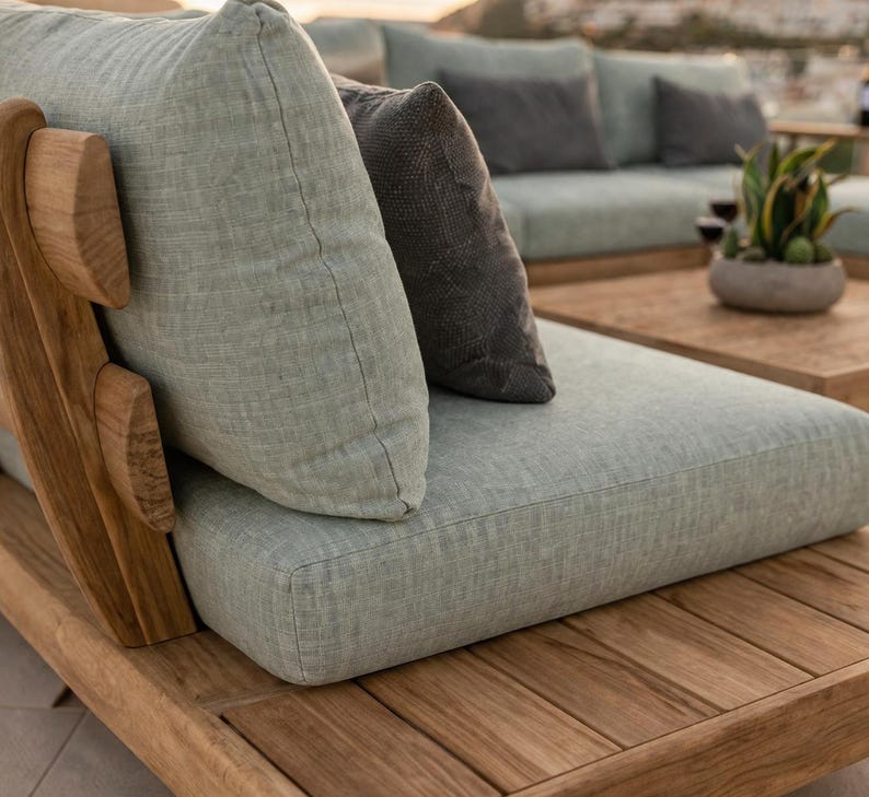 May include: A modular outdoor sofa section constructed from natural teak wood with a slatted base and frame. It features a thick, light sage green fabric seat cushion and a matching back cushion, complemented by a dark charcoal gray accent pillow.