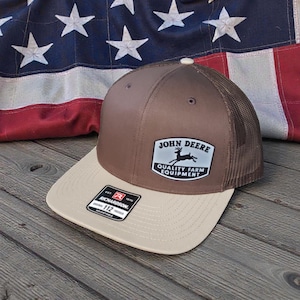 May include: A brown and tan baseball cap with a mesh back. The cap features a patch with the text "JOHN DEERE QUALITY FARM EQUIPMENT" and a deer logo. The cap is set against an American flag.