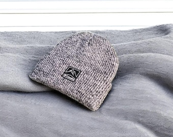 May include: A gray and black knit beanie with a small black patch that says "Columbia" on it. The beanie is sitting on a gray blanket.