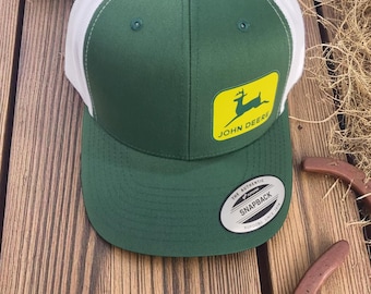 Peut inclure: Casquette snapback verte et blanche avec le logo John Deere. Le chapeau a un panneau avant et une visière verts, avec un dos en mesh blanc. Le logo John Deere est un rectangle jaune avec un cerf vert. Fermeture à pression.