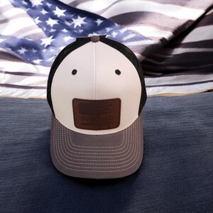 May include: A white and brown baseball cap with a black mesh back. The cap has a brown leather patch with a small American flag design on the front. The cap is sitting on a blue and white striped fabric.