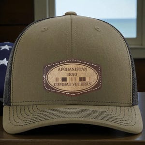 May include: Olive green and black mesh-backed baseball cap. The cap features a leather patch with the words "AFGHANISTAN IRAQ COMBAT VETERAN" in a brown oval. The cap has a curved brim and a button on top.