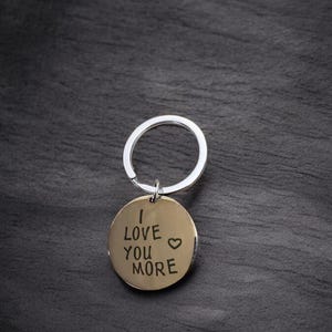 May include: A gold-colored keychain with a silver ring. The keychain has a round tag with the words "I love you more" engraved on it, along with a small heart symbol.