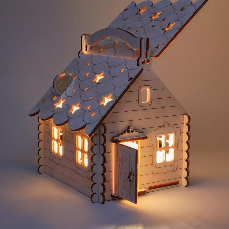 Pode incluir: Uma l&acirc;mpada em forma de casa de madeira com telhado inclinado e porta aberta. O telhado apresenta recortes em forma de estrela que emitem luz. As janelas e a porta tamb&eacute;m brilham, criando um ambiente acolhedor. A casa &eacute; feita de madeira clara.