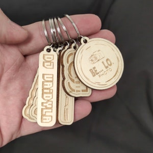 Pode incluir: Um conjunto de cinco chaveiros de madeira com designs diferentes. Um chaveiro é redondo e apresenta o texto "BE...LO. PHOTO". Outro chaveiro é retangular e apresenta o texto "DJ VAIDYALA". Os chaveiros estão presos a argolas de chave prateadas.