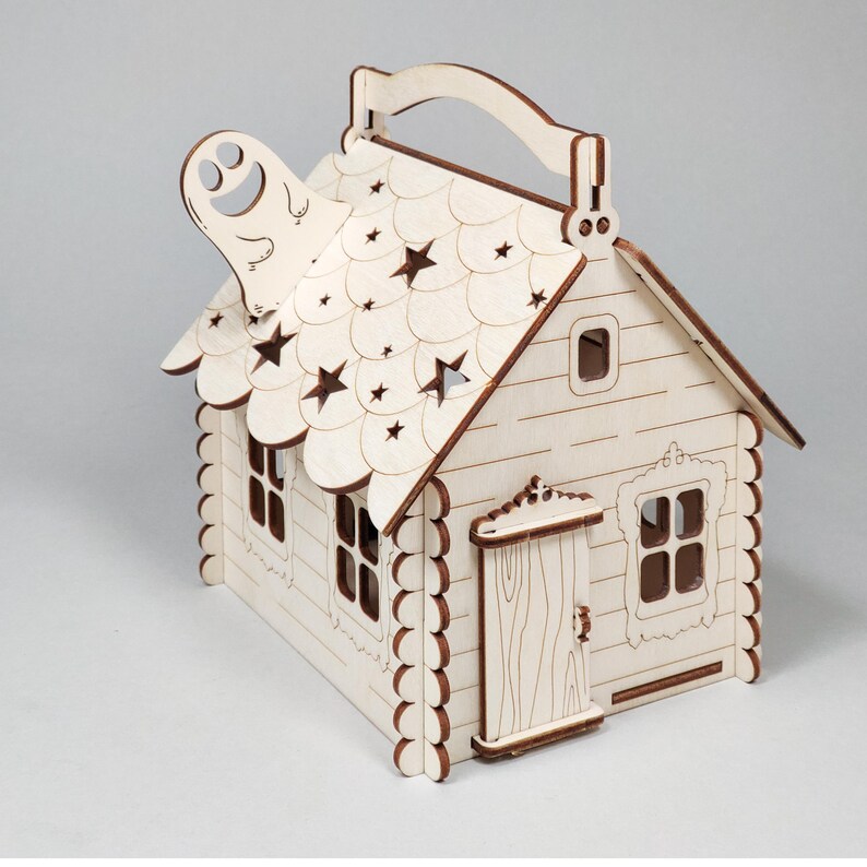 Pode incluir: Uma caixa de madeira em forma de casa com uma al&ccedil;a, cortada de madeira clara. O telhado tem recortes de estrelas e um recorte em forma de fantasma. A casa tem janelas, uma porta e acabamentos decorativos.