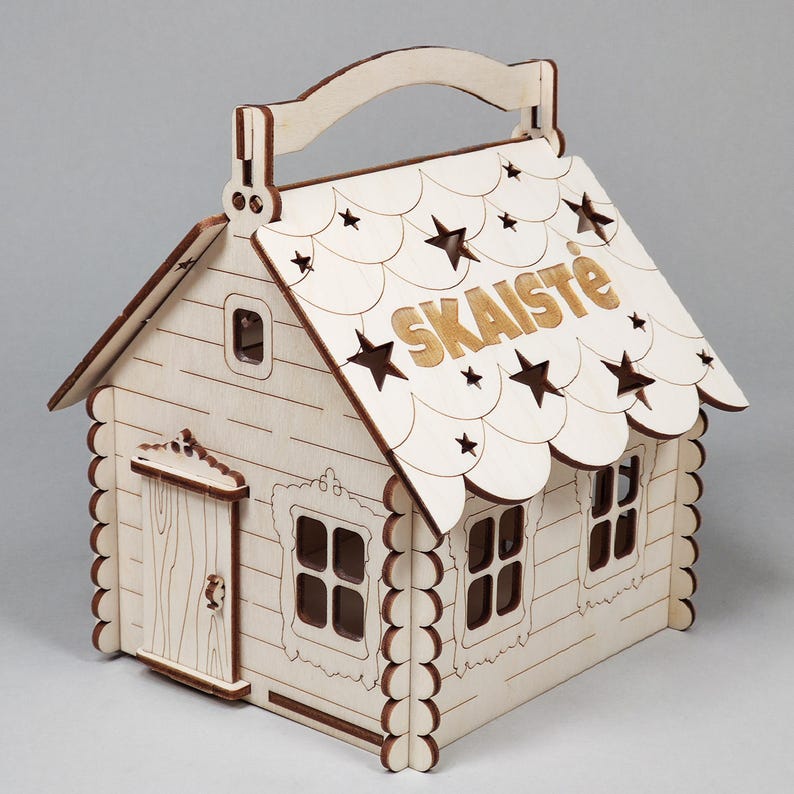 Pode incluir: Uma caixa de madeira em forma de casa com uma al&ccedil;a, janelas e uma porta. O telhado apresenta recortes de estrelas e o nome "SKAISTĖ" em letras douradas. A casa &eacute; feita de madeira clara com um acabamento natural.