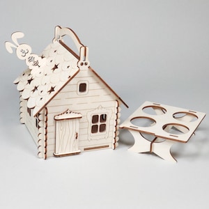 Pode incluir: Um item decorativo em forma de casa de madeira com uma al&ccedil;a e um coelho no telhado. A casa tem uma porta, janelas e um telhado com padr&atilde;o de estrela. Um suporte de madeira separado com quatro recortes circulares est&aacute; ao lado da casa.