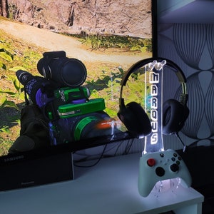 Pode incluir: Um suporte de auscultadores para jogos branco com uma base de acrílico transparente e uma luz LED branca. O suporte tem a palavra "MARQUIS" impressa. Os auscultadores são pretos com um microfone branco. O suporte está sobre uma mesa ao lado de um comando de consola de jogos.