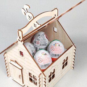 Pode incluir: Uma cesta em forma de casa de madeira com uma al&ccedil;a e um recorte de coelho no topo. Cont&eacute;m tr&ecirc;s ovos Kinder Surpresa. A casa tem janelas e uma porta, todas recortadas em madeira. Os ovos s&atilde;o brancos com embalagens coloridas.