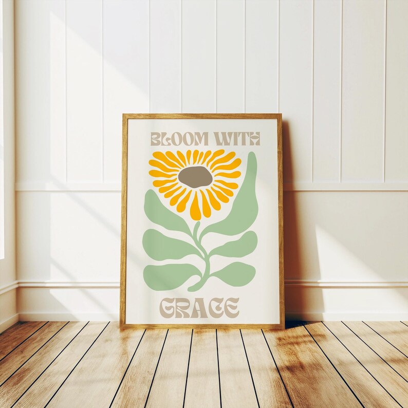 May include: A framed art print featuring a stylized sunflower with yellow petals and a brown center, green leaves, and the words "Bloom with Grace" in a retro font. The artwork is set against a white background.