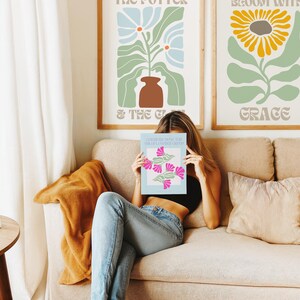 May include: A woman sits on a beige sofa, holding a book with pink floral illustrations. Two framed botanical art prints hang on the wall behind her. One features a blue flower, the other a yellow sunflower. A tan throw blanket and a striped pillow are also visible.