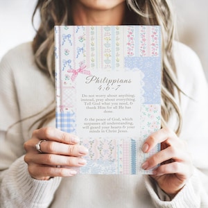 May include: A notebook with a patchwork design cover featuring floral patterns in pastel pinks, blues, and whites. The cover includes the text "Philippians 4:6-7". The notebook is held by a person.
