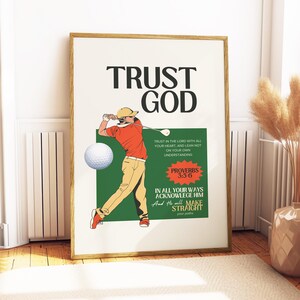 Op de afbeelding: Een ingelijste print met de woorden "TRUST GOD" in grote zwarte letters. De afbeelding toont een golfer in een oranje shirt en kaki broek die een golfclub zwaait. De print bevat een groene box met tekst uit Spreuken 3:5-6.