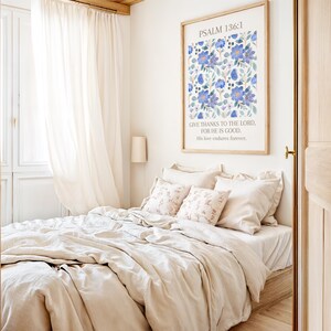 Op de afbeelding: Een slaapkamer met een bed met beige beddengoed en decoratieve kussens. Een ingelijste print met blauwe bloemenkunst en de tekst van Psalm 136:1 hangt boven het bed. Transparante witte gordijnen omlijsten een raam. De kamer heeft een warme sfeer.