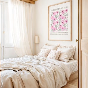 May include: A bedroom with a bed dressed in beige bedding and patterned pillows. A framed print with pink floral art and text is mounted on the wall. Sheer curtains and a lamp are also visible.