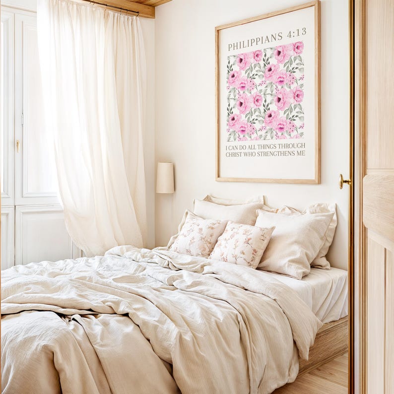 May include: A bedroom with a bed dressed in beige bedding and patterned pillows. A framed print with pink floral art and text is mounted on the wall. Sheer curtains and a lamp are also visible.