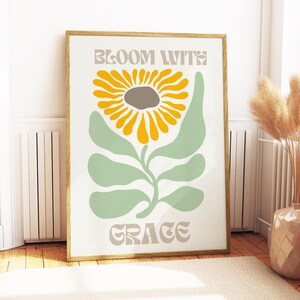 May include: A framed art print featuring a stylized sunflower with yellow petals and a gray center, set against a light green stem and leaves. The words "BLOOM WITH GRACE" are printed in a muted beige font. The artwork is displayed in a room with a beige rug and wooden floor.