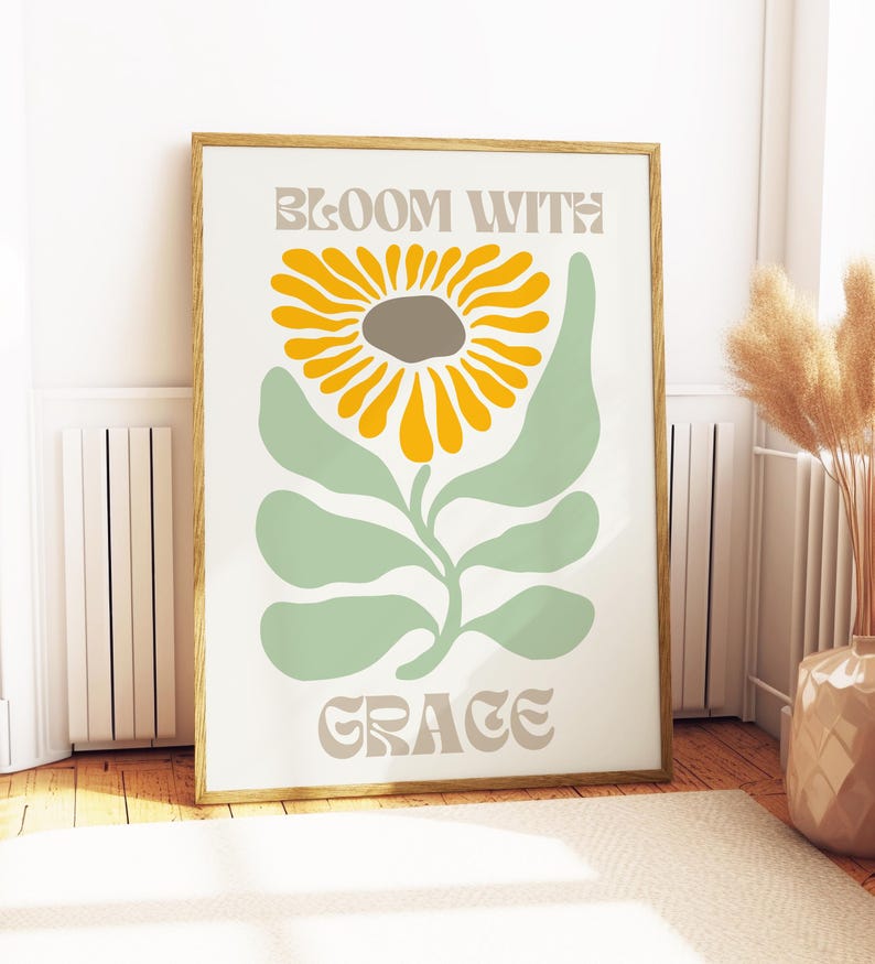 May include: A framed art print featuring a stylized sunflower with yellow petals and a gray center, set against a light green stem and leaves. The words "BLOOM WITH GRACE" are printed in a muted beige font. The artwork is displayed in a room with a beige rug and wooden floor.