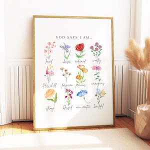 May include: A framed art print with a gold-colored frame. The print features watercolor illustrations of various flowers and the text "GOD SAYS I AM..." followed by words like "loved," "chosen," and "redeemed." The artwork is displayed on a white background.